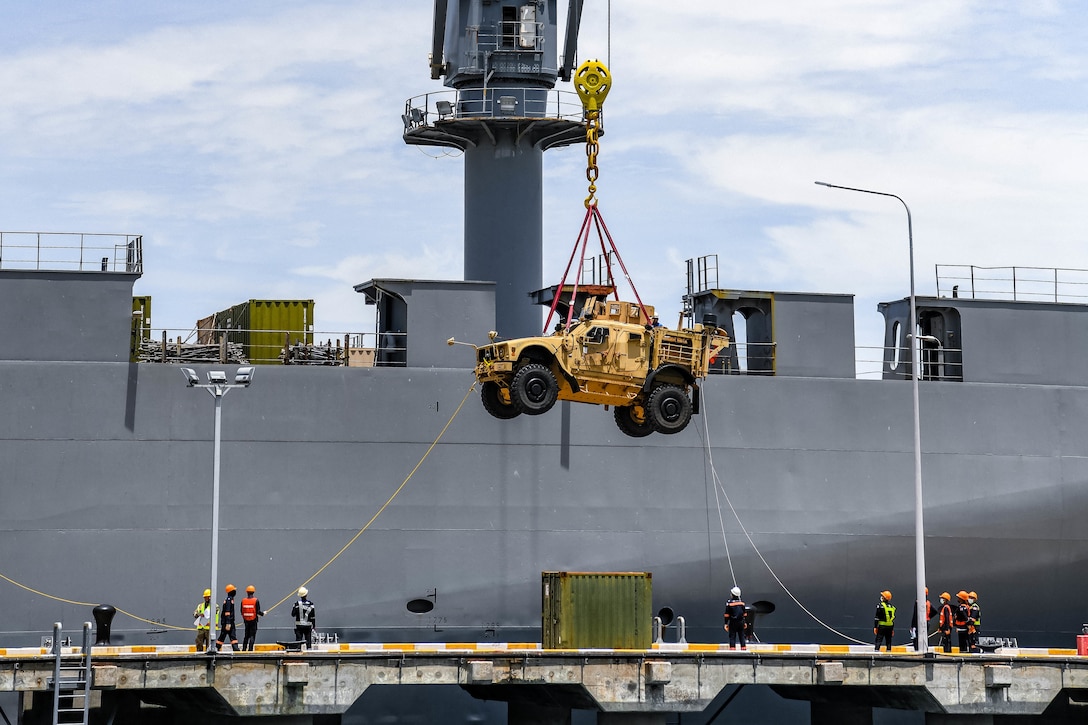 A large military vehicle is lowered by cable from a ship to a dock.