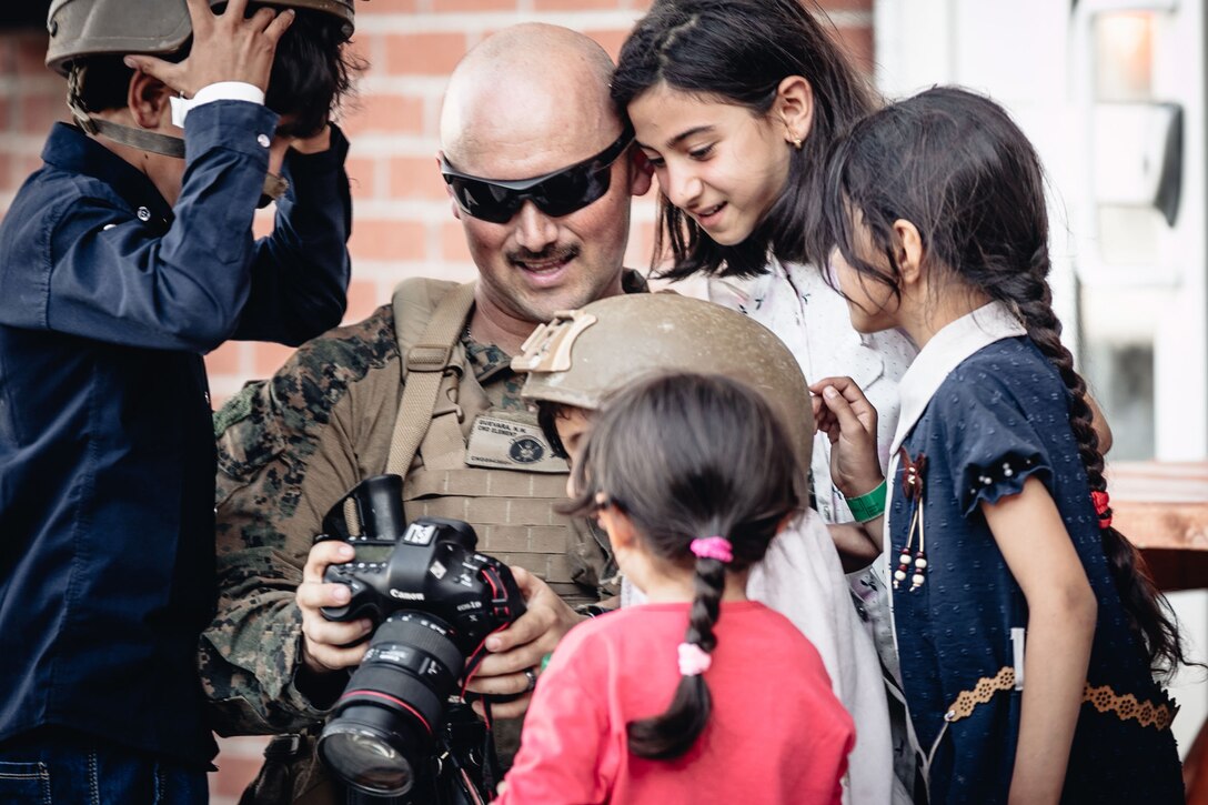 A Marine shows a camera to a group of smiling children.