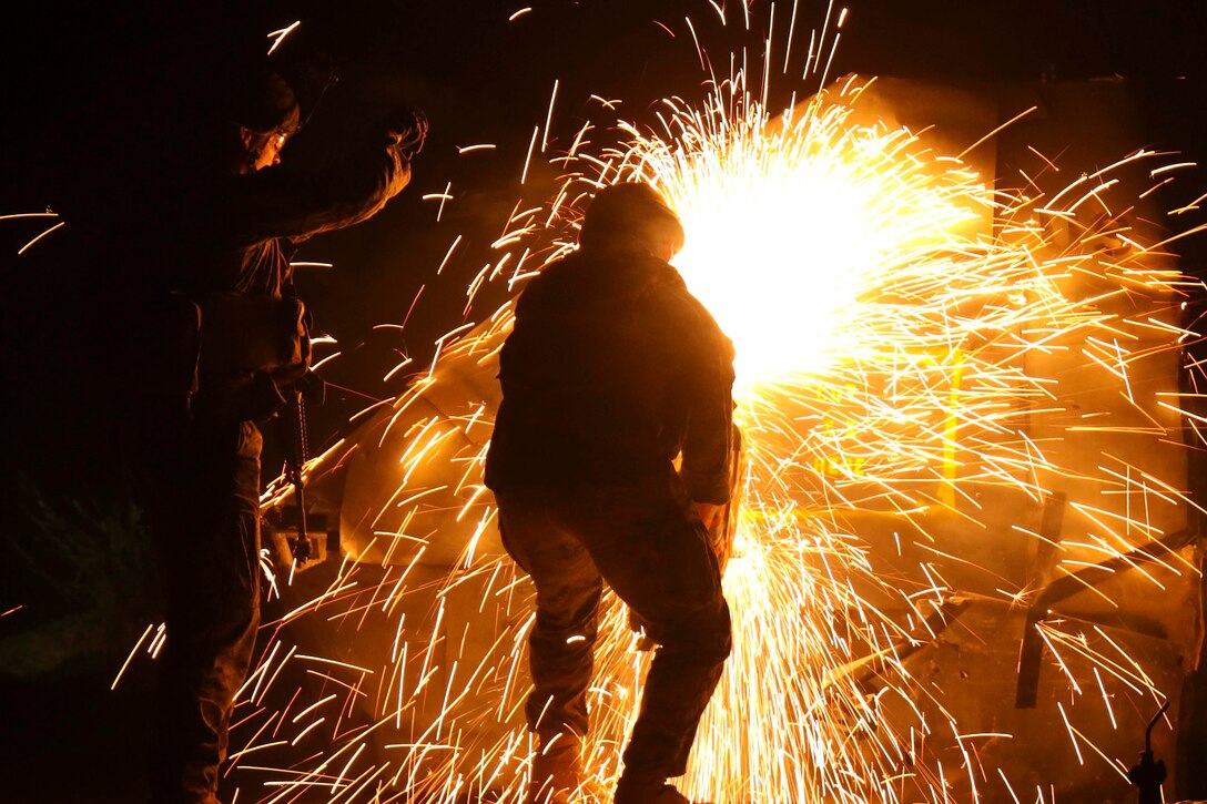 Two Marines use a saw in a dark room as sparks fly around.