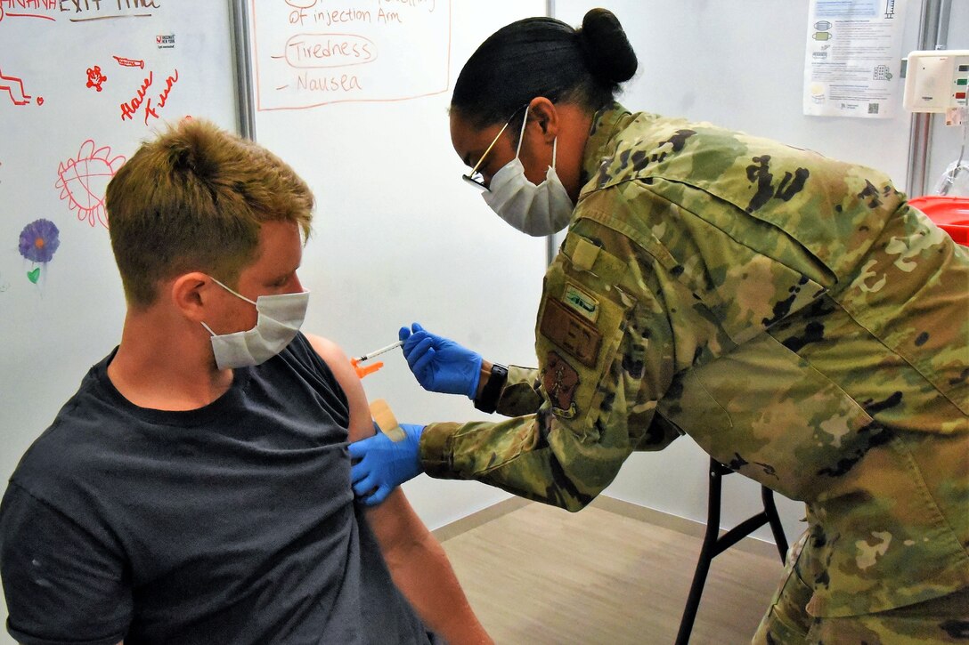 An airman wearing a face mask and gloves holding a syringe leans over to give a soldier wearing a face mask a vaccination.