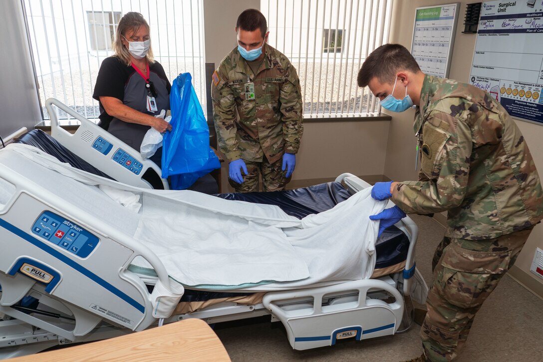 A nursing assistant wearing a face mask and holding plastic bags teaches cleaning protocols to two soldiers wearing face masks and gloves while they put a sheet on a hospital bed.