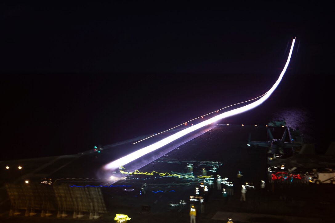Sailors stand on the deck of a ship as an aircraft takes off illuminated by different colors.