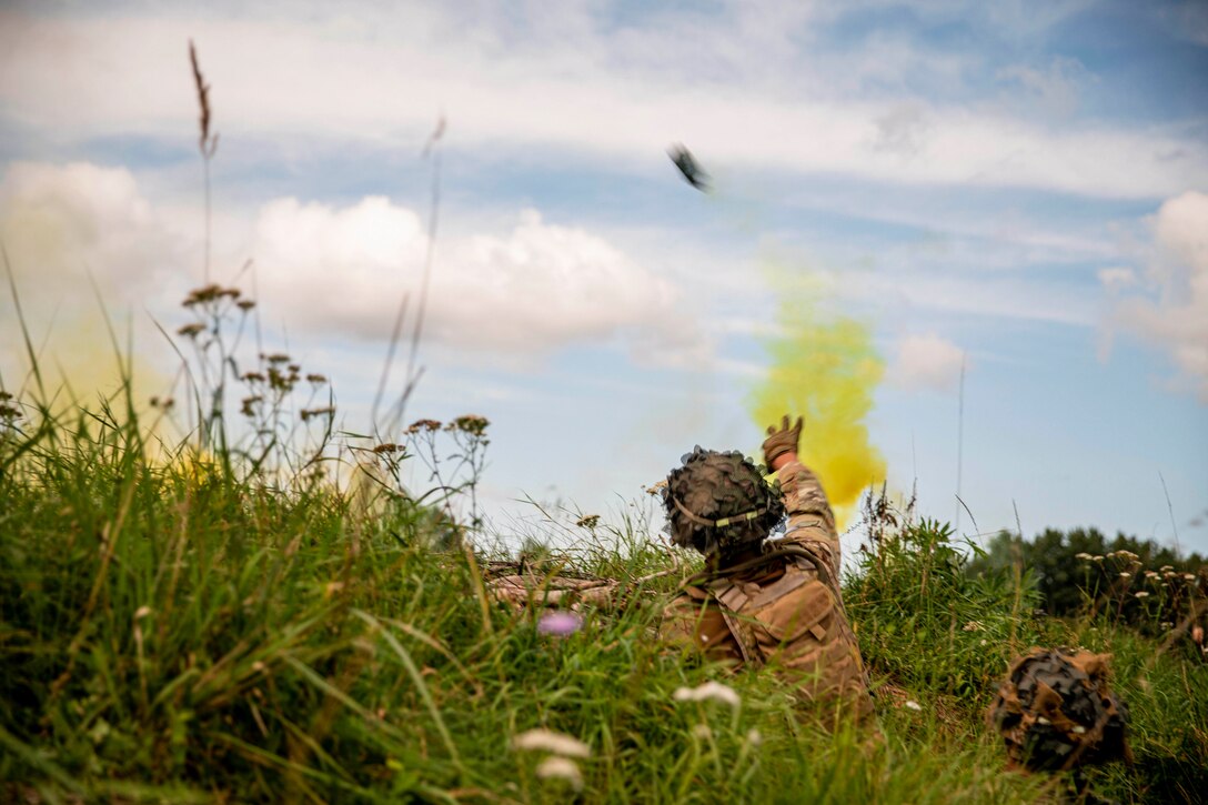 A National Guardsman throws a smoke grenade while lying in the grass.