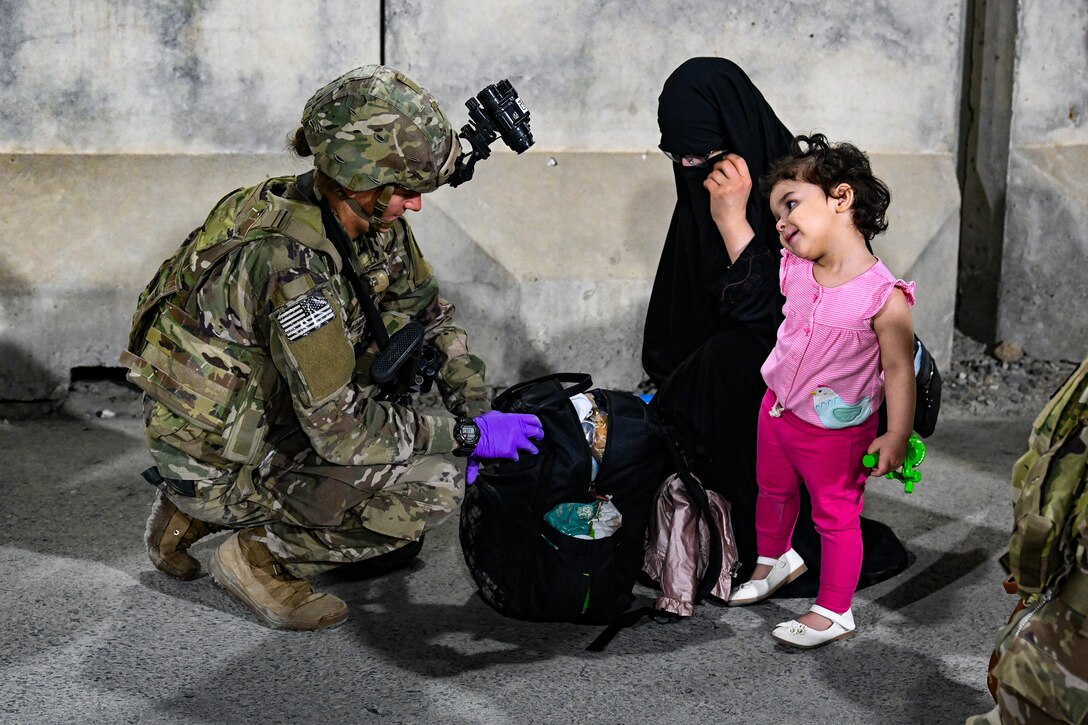A soldier crouches on the ground and inspects the contents of a bag. A small girl and her mother watch.