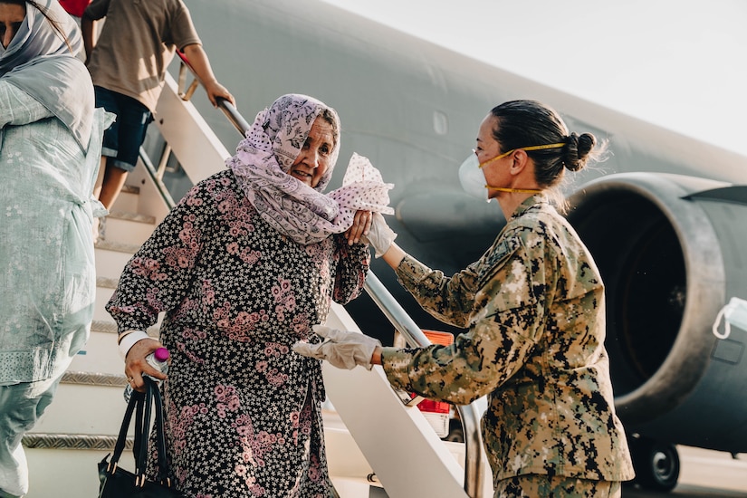 A woman in a military uniform assists an older woman down the stairs of a military aircraft.