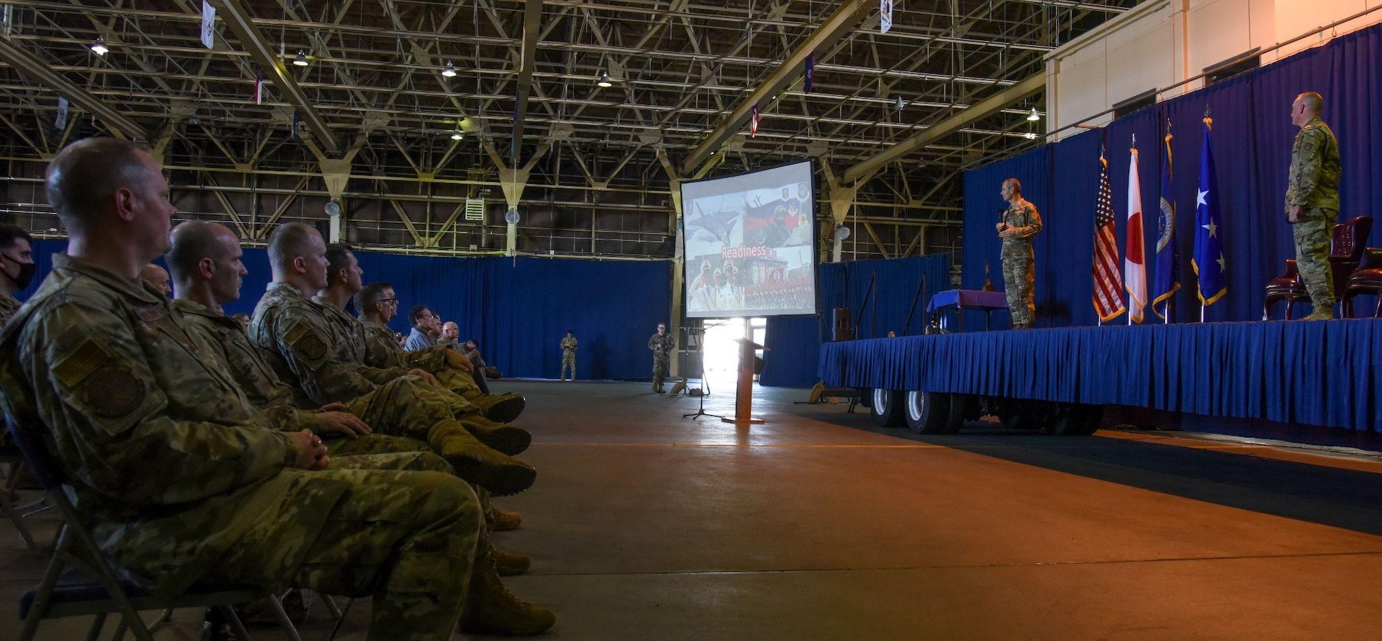 Service members listening to another service member  speaking on a stage.