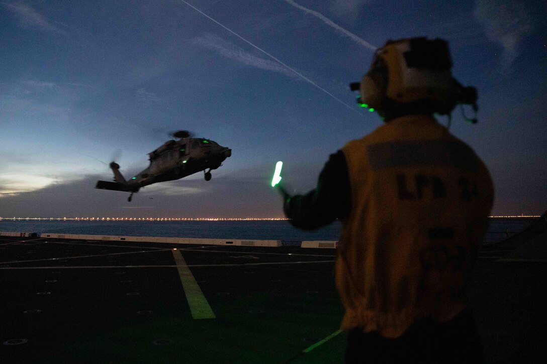 A man directs a helicopter hovering above the flight deck of a ship.