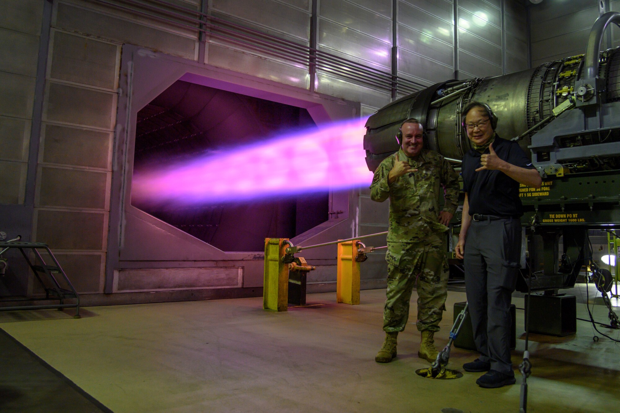 Two people stand beside running jet engine emitting blueish, purple flames.