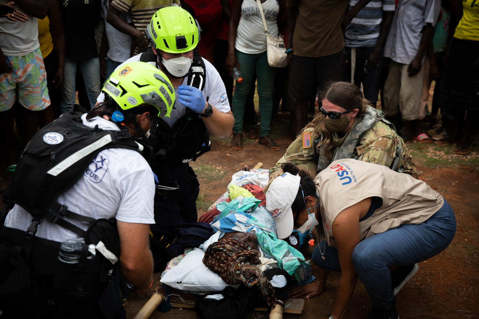 A service member and several civilians tend to a person in a stretcher on the ground.