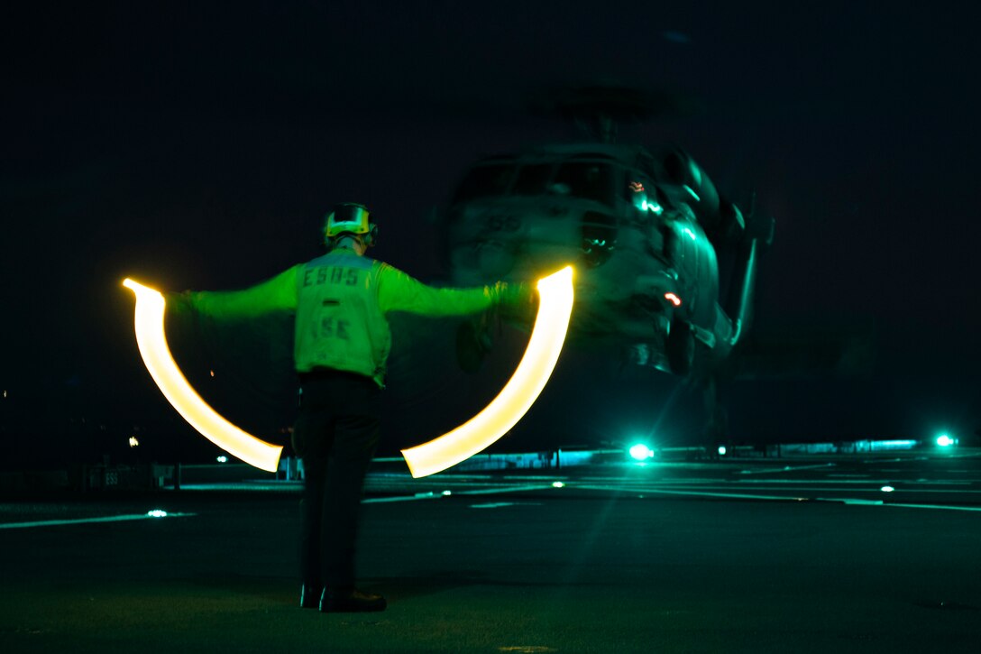 An airman uses lights to help a helicopter land on a ship at night.