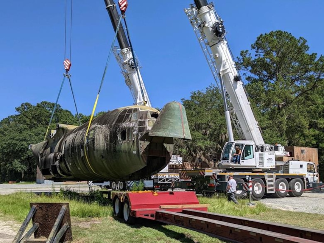 A C-130 fuselage is being placed onto a flatbed truck for transport.