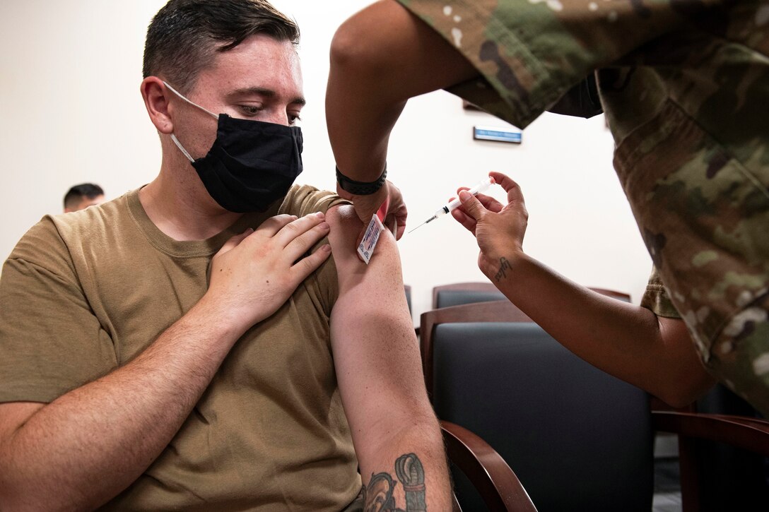 A man bending over and holding a syringe gives a COVID-19 vaccine to an airman wearing a face mask while seated.