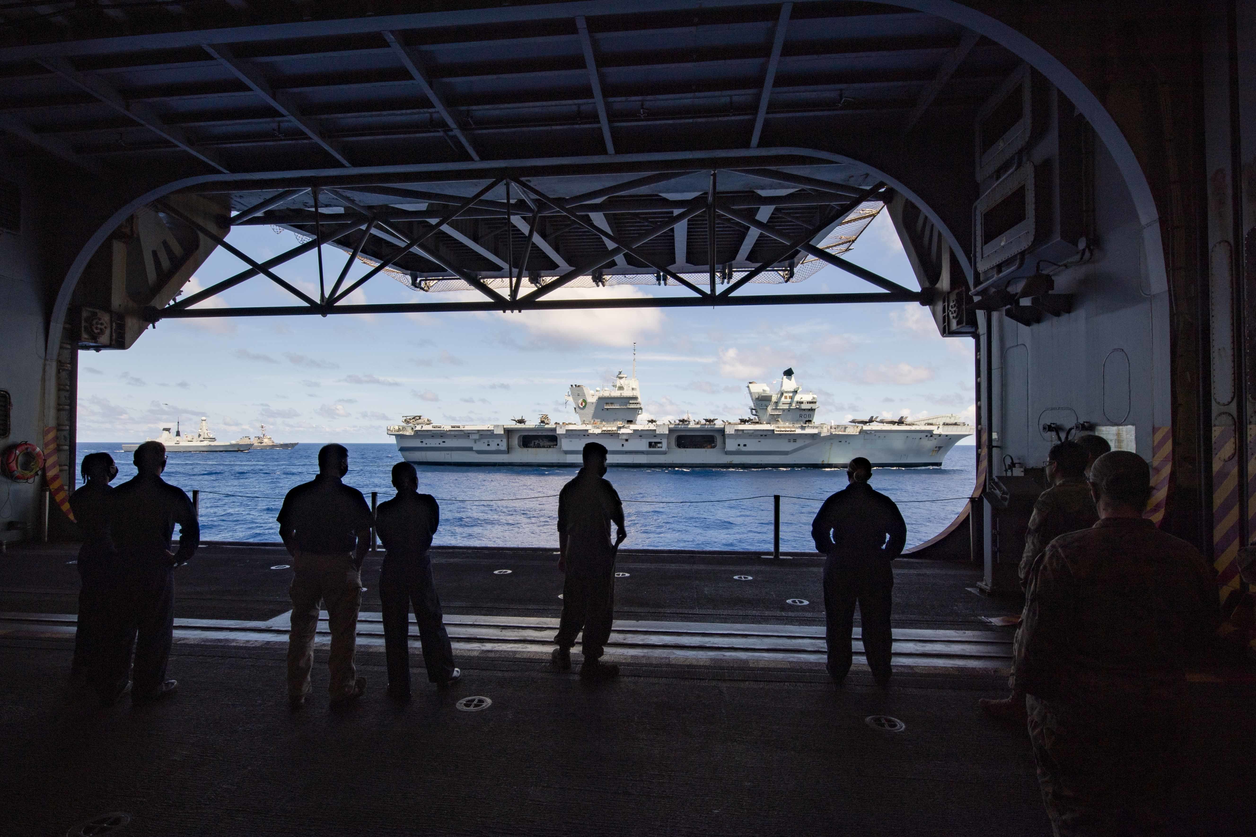 Sailors and Marines watch the Royal Navy aircraft carrier HMS Queen Elizabeth (R 08), foreground, followed by the Royal Navy destroyer HMS Defender (D 36) in the ship�s hangar bay