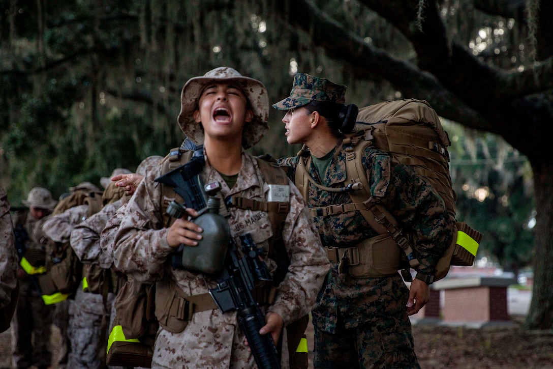 A Marine Corps recruit shouts while carrying gear under a tree while fellow recruits stand in a line.