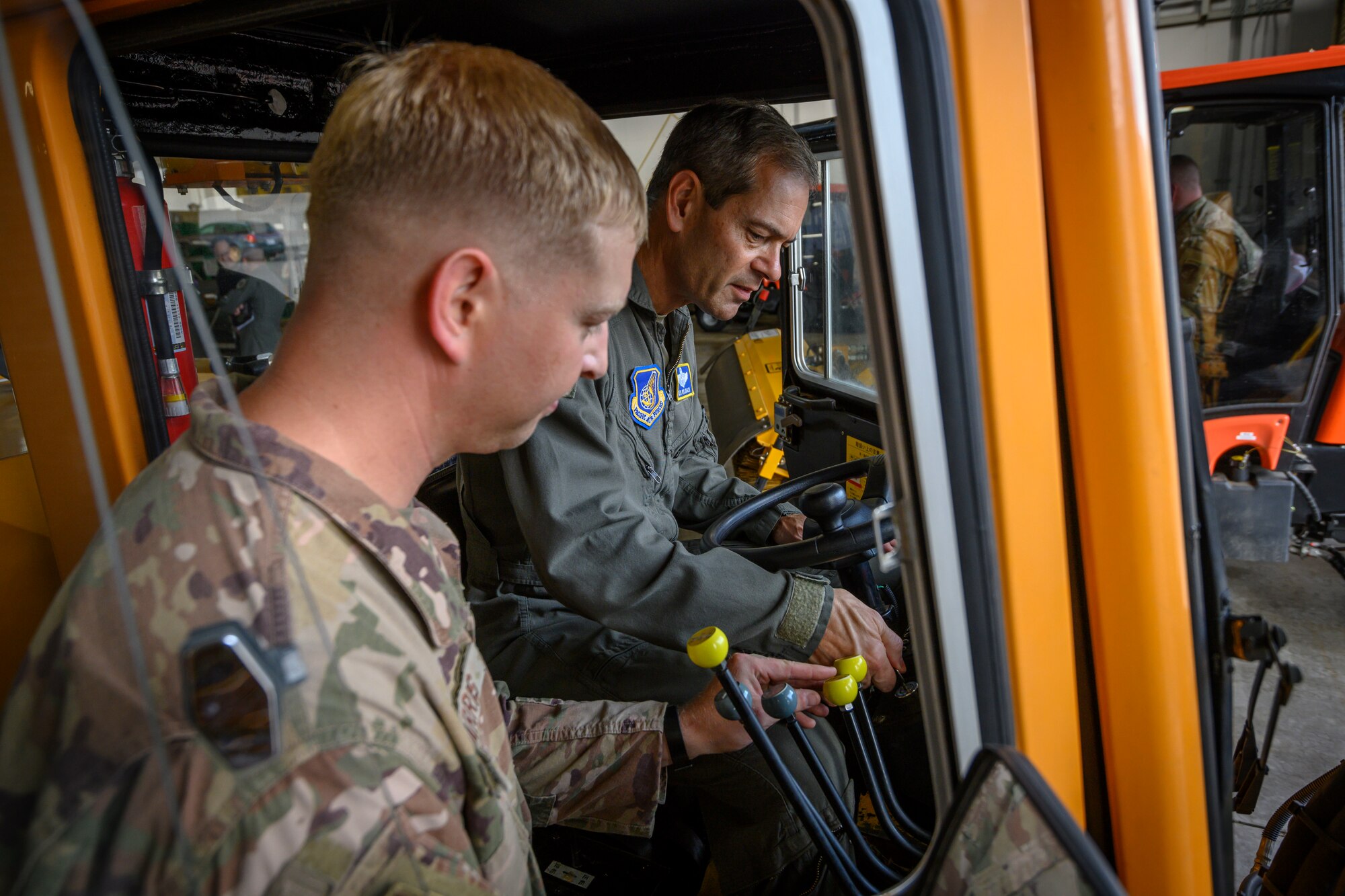 A military member in a heavy operating vehicle is being instructed by another military member.