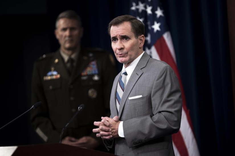 A man in a suit speaks at a podium; an army officer stands in the background next to an American flag.