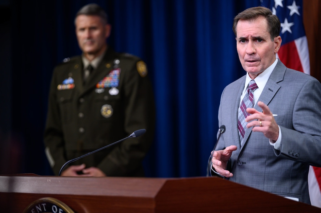 A man stands at a lectern as another man stands behind.