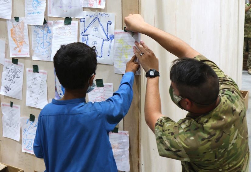 A U.S. Army Central Soldier helps an Afghan evacuee boy display his art work with others at Camp Buehring, Kuwait, Aug. 25, 2021. Soldiers joined in activities and spent time with the children as their families went through arrival processes at the facility. (U.S. Army photo by Pfc. Katelynn Bissell)
