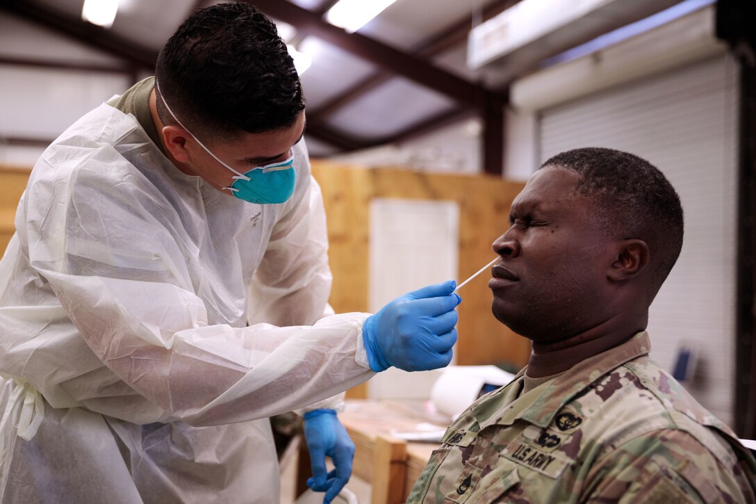 A soldier holding a nasal swab and wearing a face mask, gloves and a medical gown bends down to swab the nose of a  soldier, who's seated.