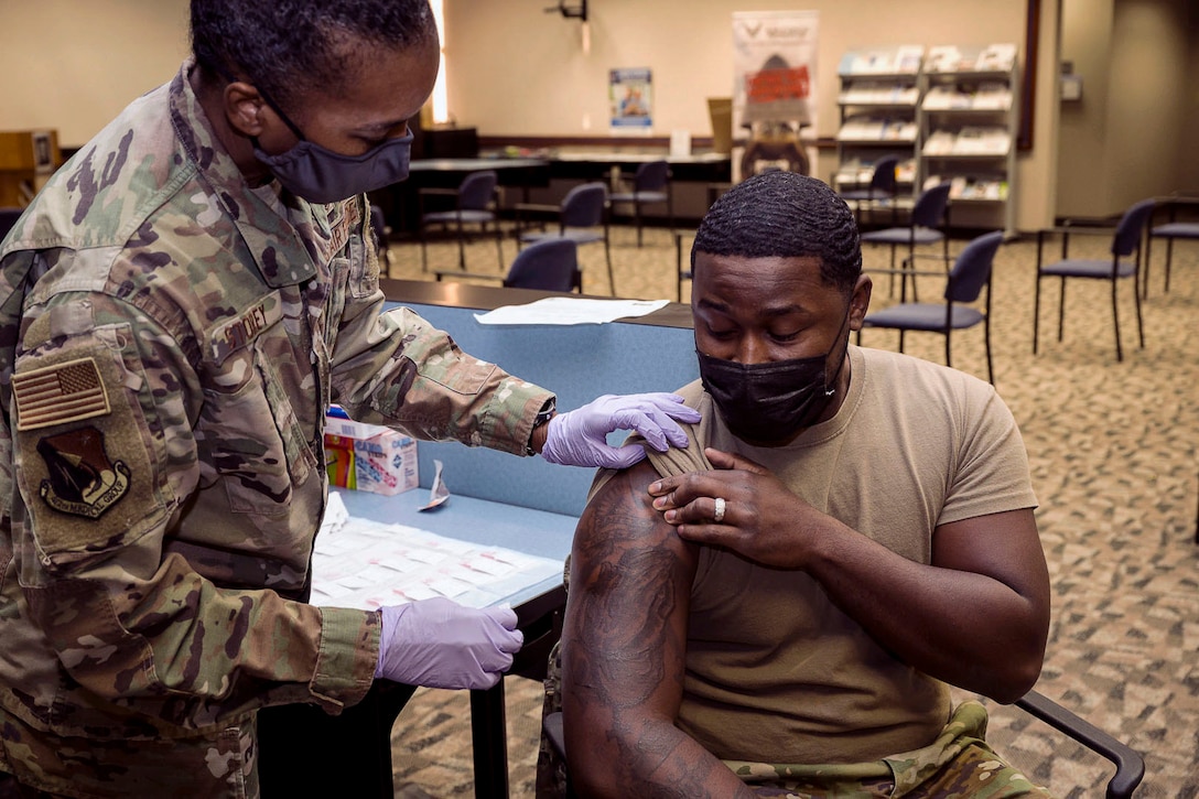 A service member, who's wearing a face mask and gloves, pulls up the right sleeve of an airman seated in a chair.