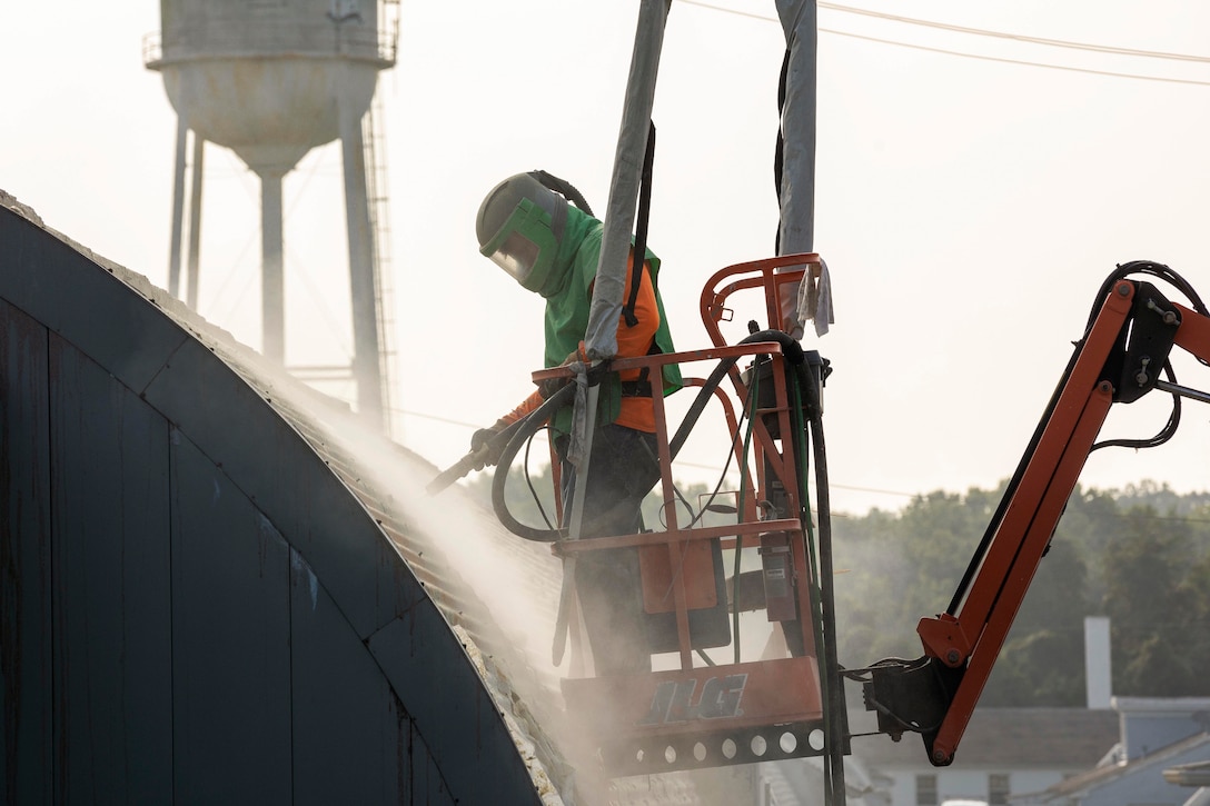 A Marine standing in a crane uses a hose to wash a building.