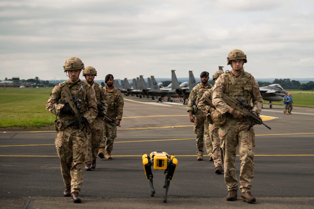 Airmen walk in two lines with a robot in between, away from a line of aircraft on a flightline.