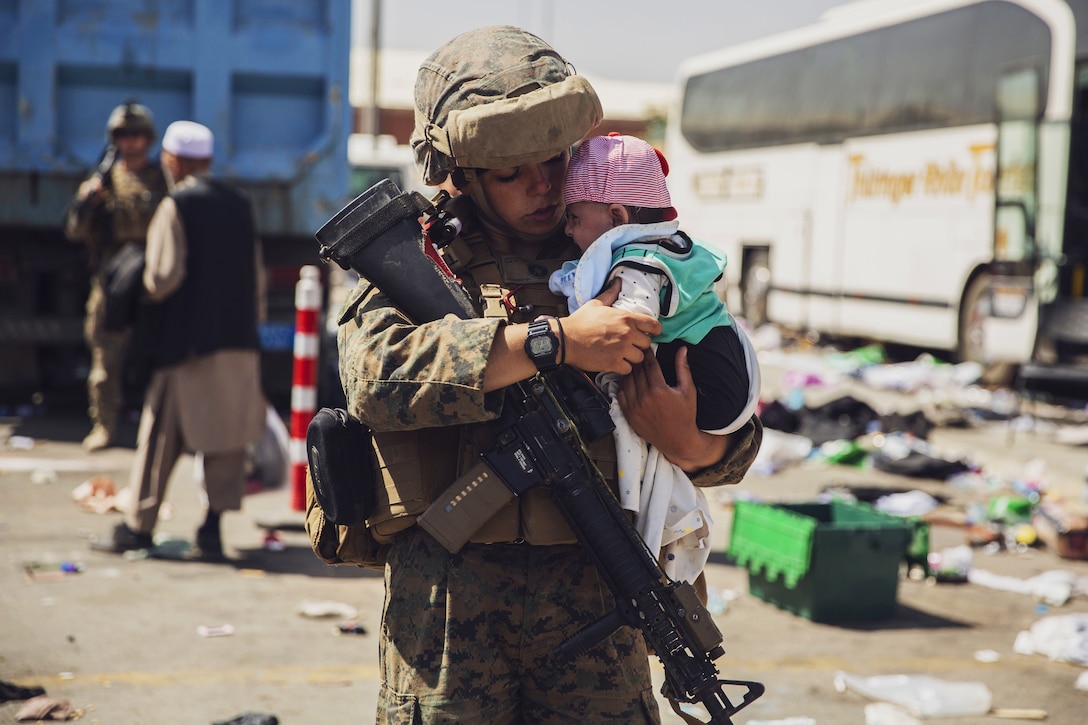A Marine holds a baby while people file in the background.