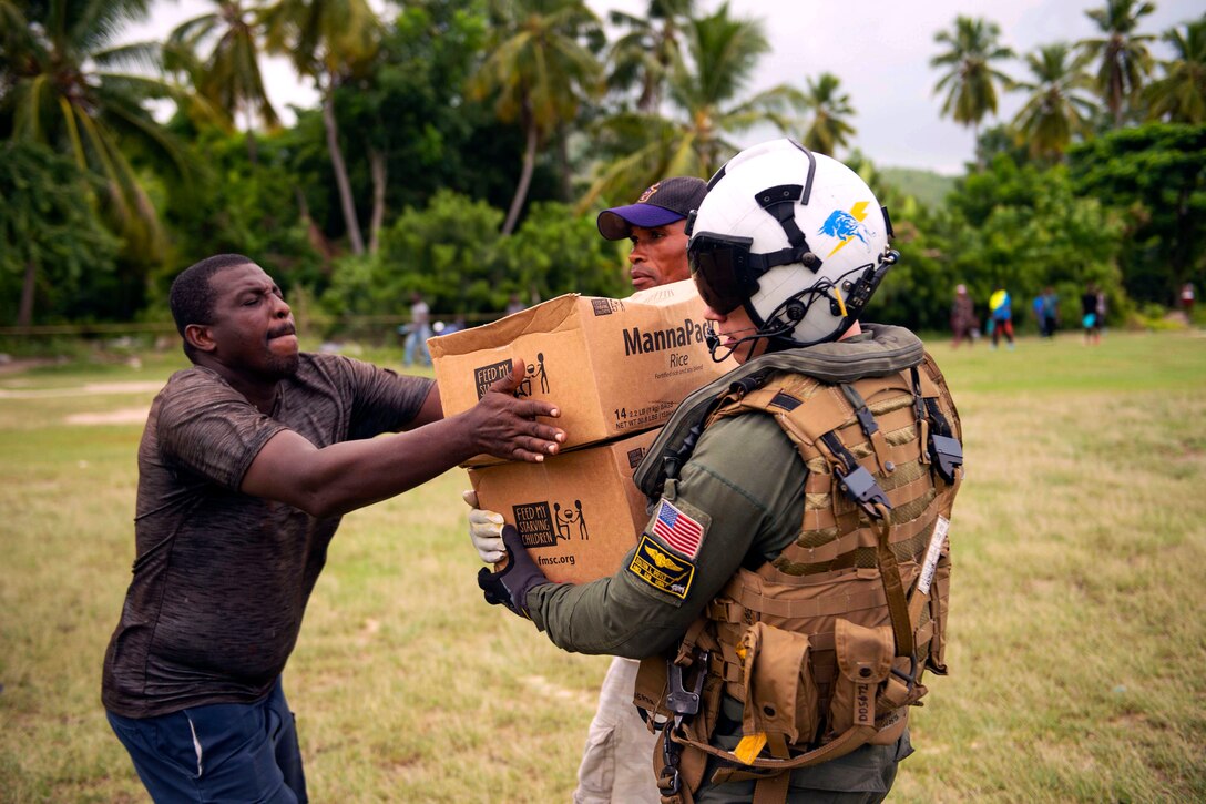 A sailor wearing a helmet passes boxes of food to a civilian in a field.