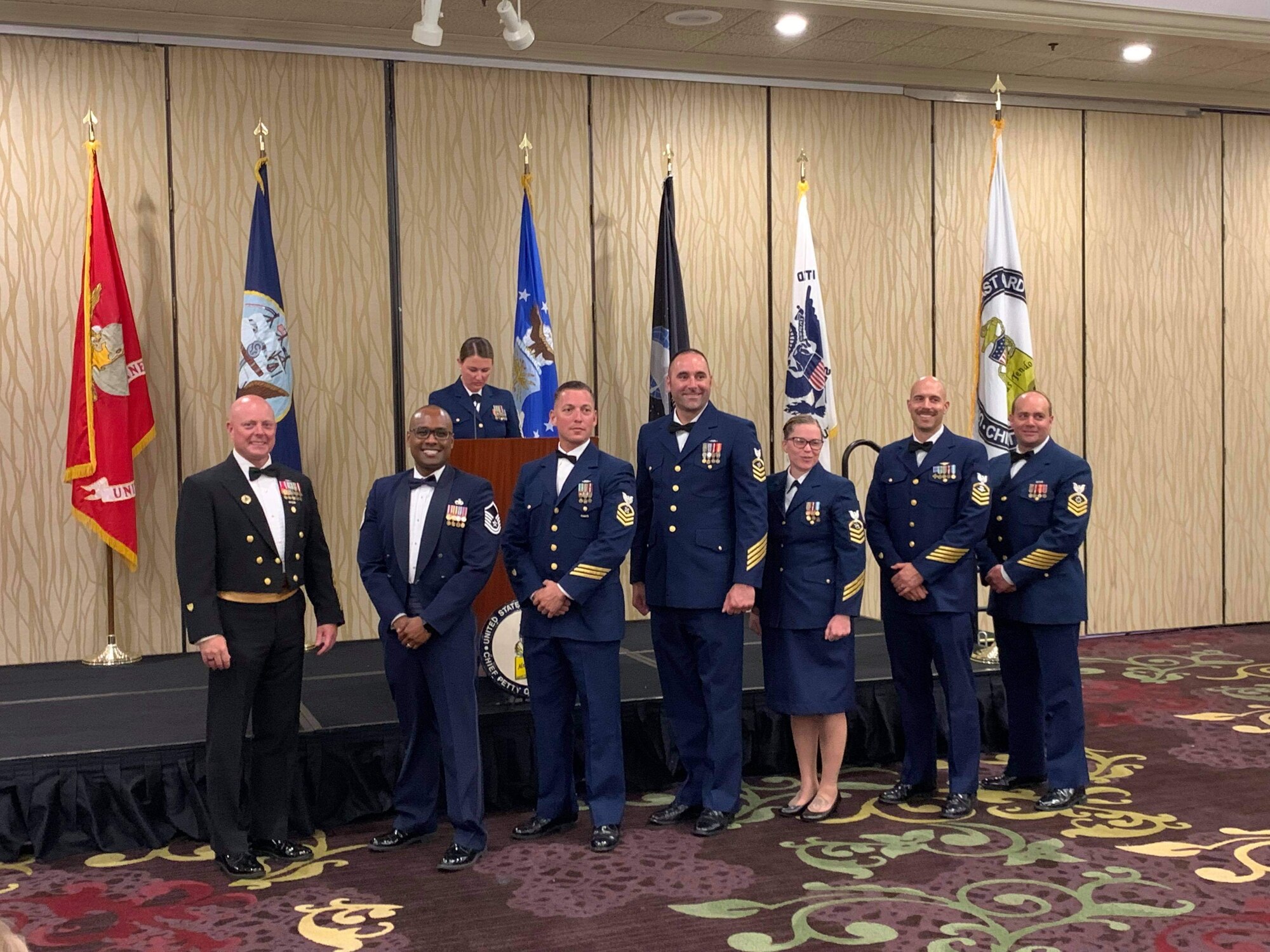 7 people stand in a room in front of flags representing every U.S. branch of service