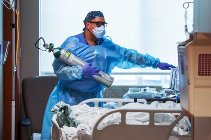 A man wearing a face mask, gloves and medical gown holds an oxygen tank.