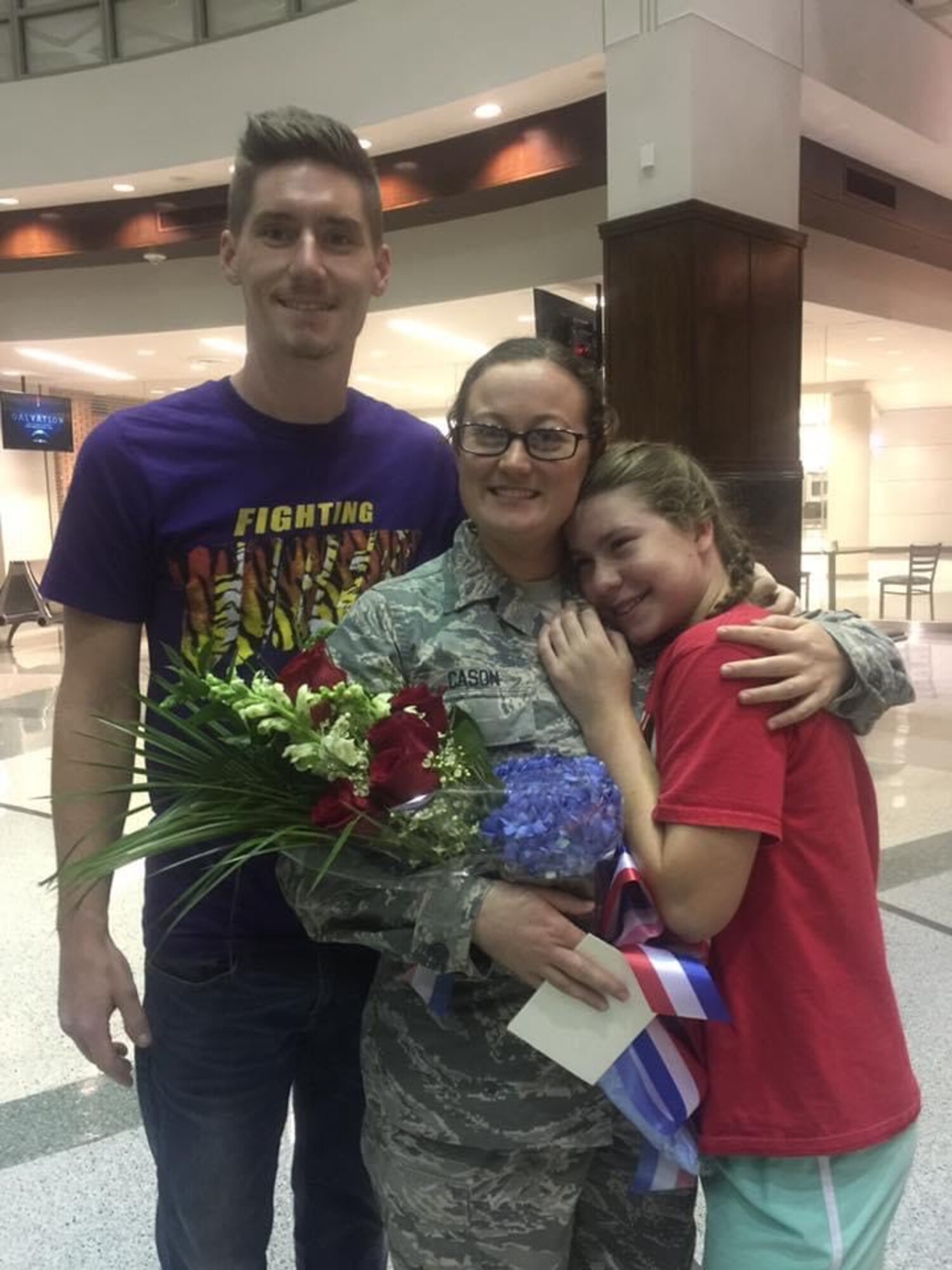 Woman holding flowers hugs young girl. Man stands near them smiling.