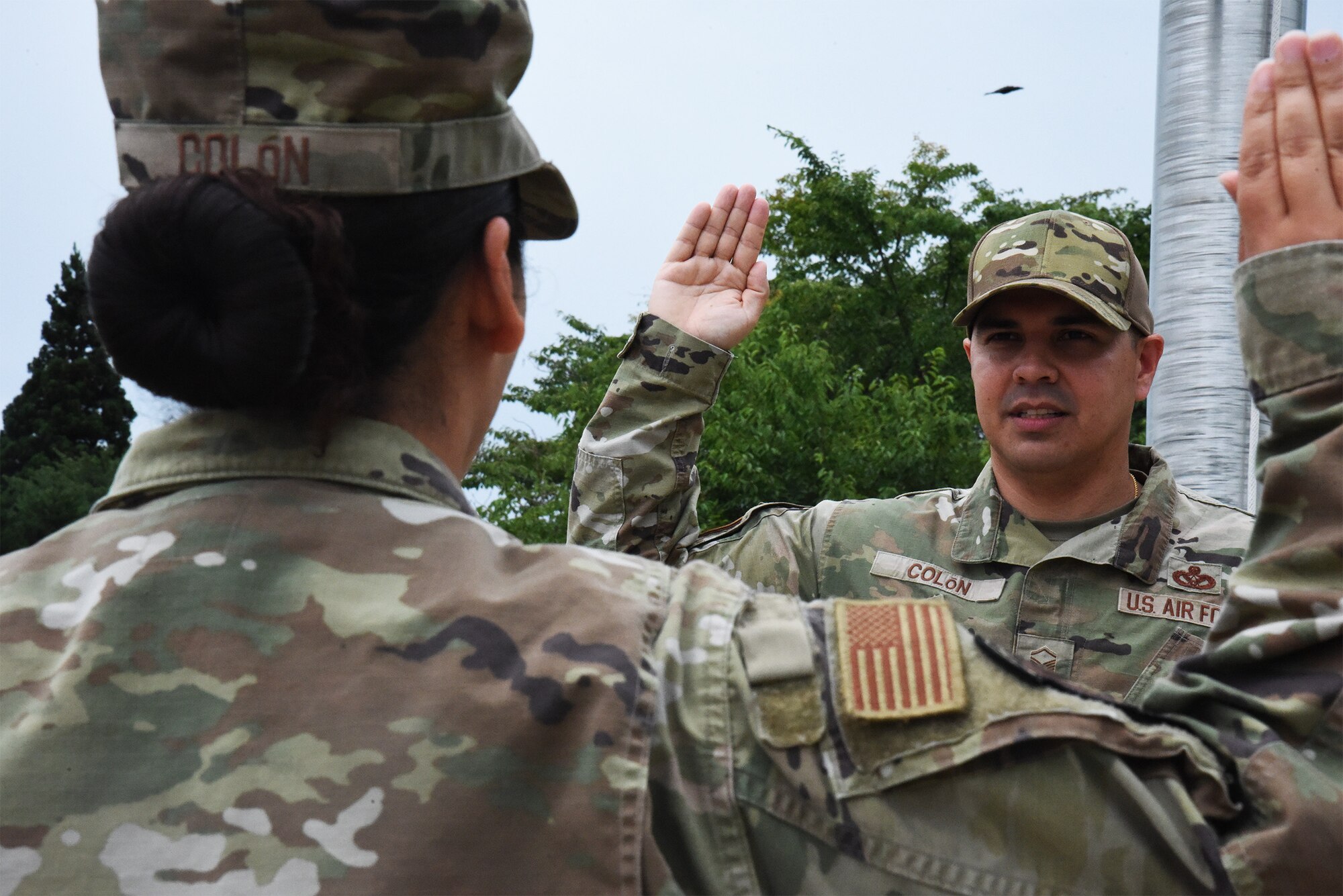 A man in uniform holds his right hand up in a square. A woman in uniform is in the foreground holding her hand up in a square.