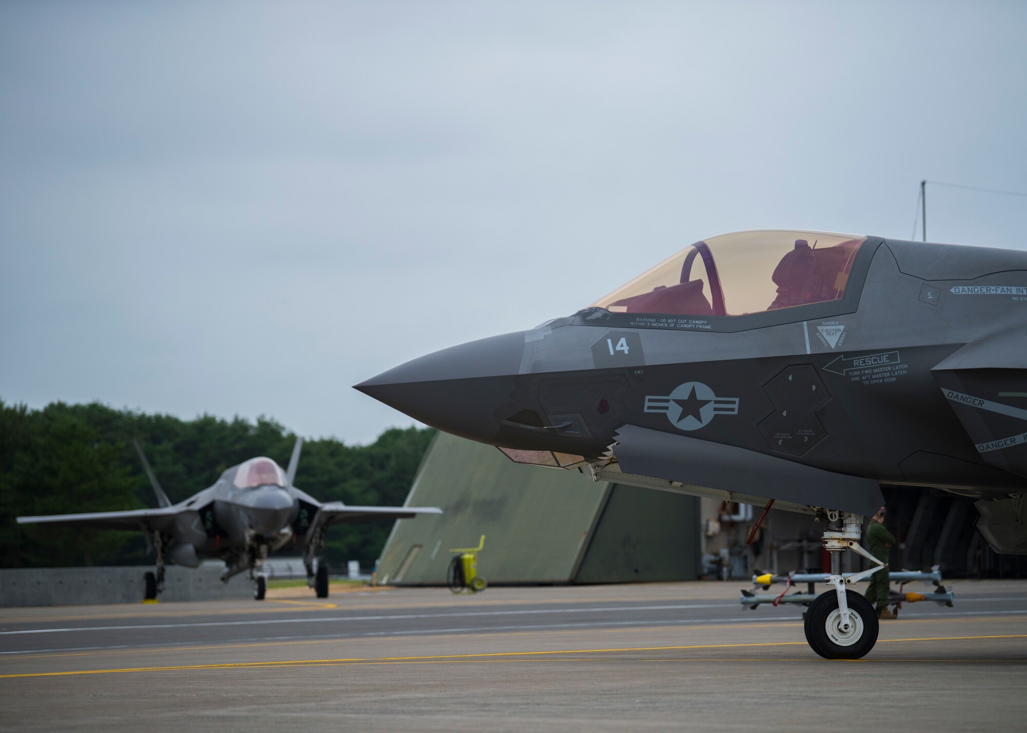 Two fighter jets sit near aircraft shelters. One close, one in the distance.
