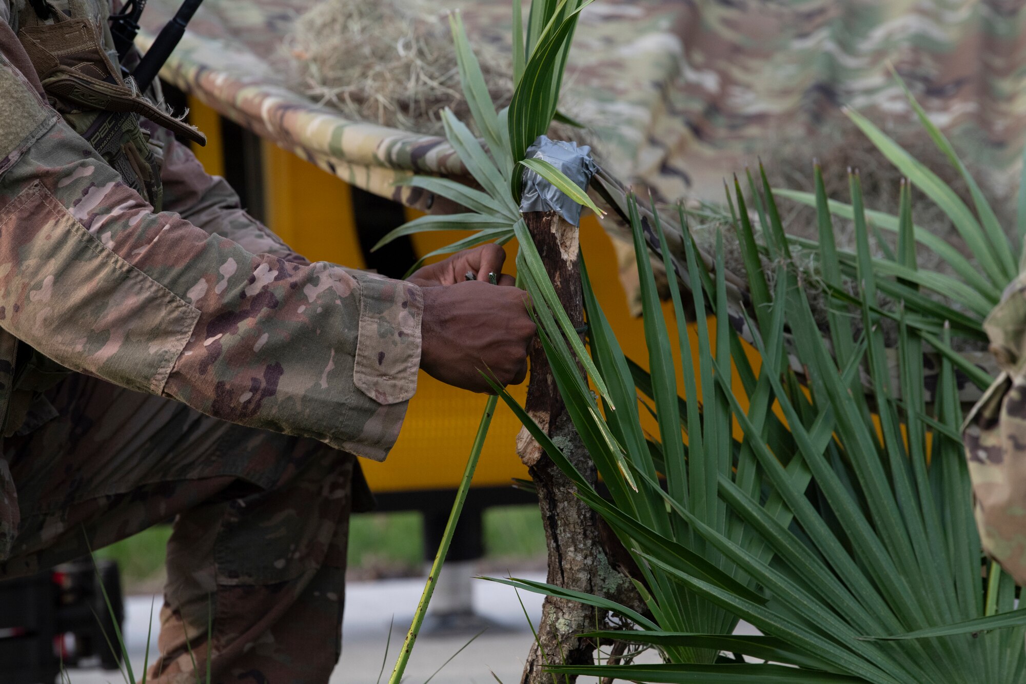 A photo of an Airman taping a palm tree leave to a stick.