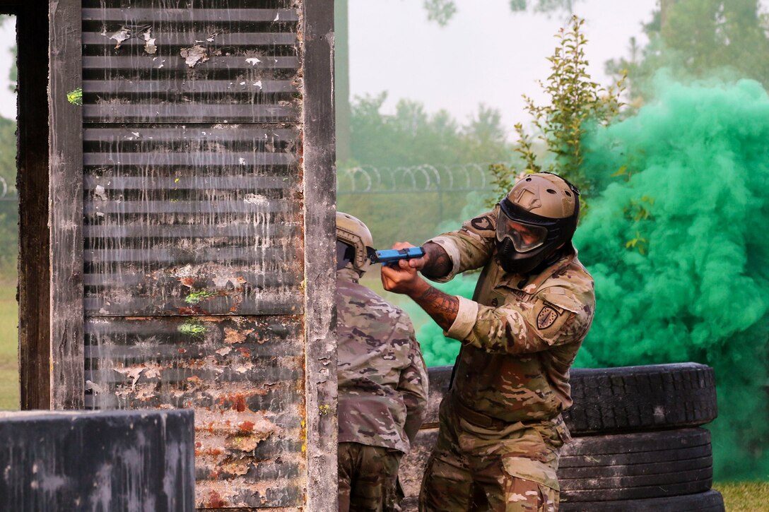 A soldier fires a weapon near a training course near fellow soldiers with clouds of green smoke in the background.