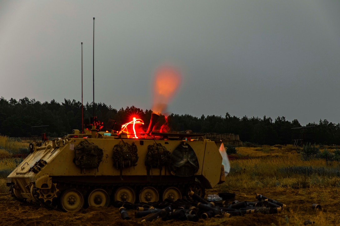 Soldier fire weapons from a tank in a field at night illuminated by red lights.