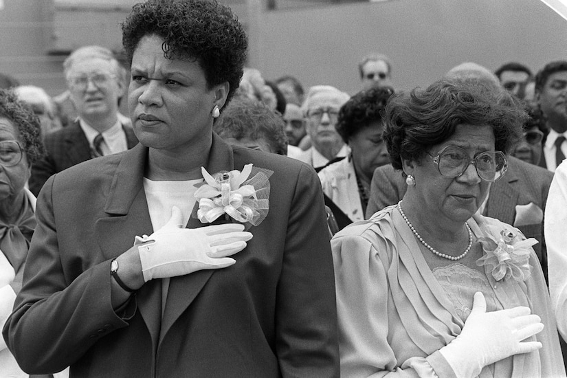 Two women put their hands over their hearts. Many people stand in the background.