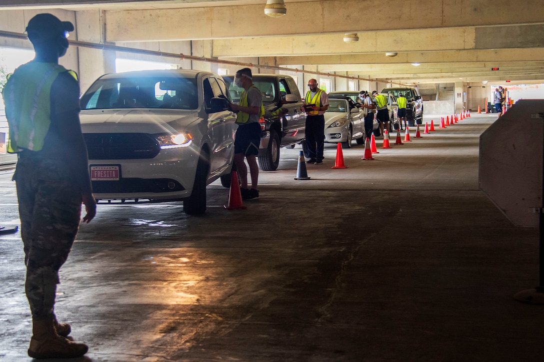 Several airmen wearing face masks screen a line of patients while still in their cars in a parking garage.