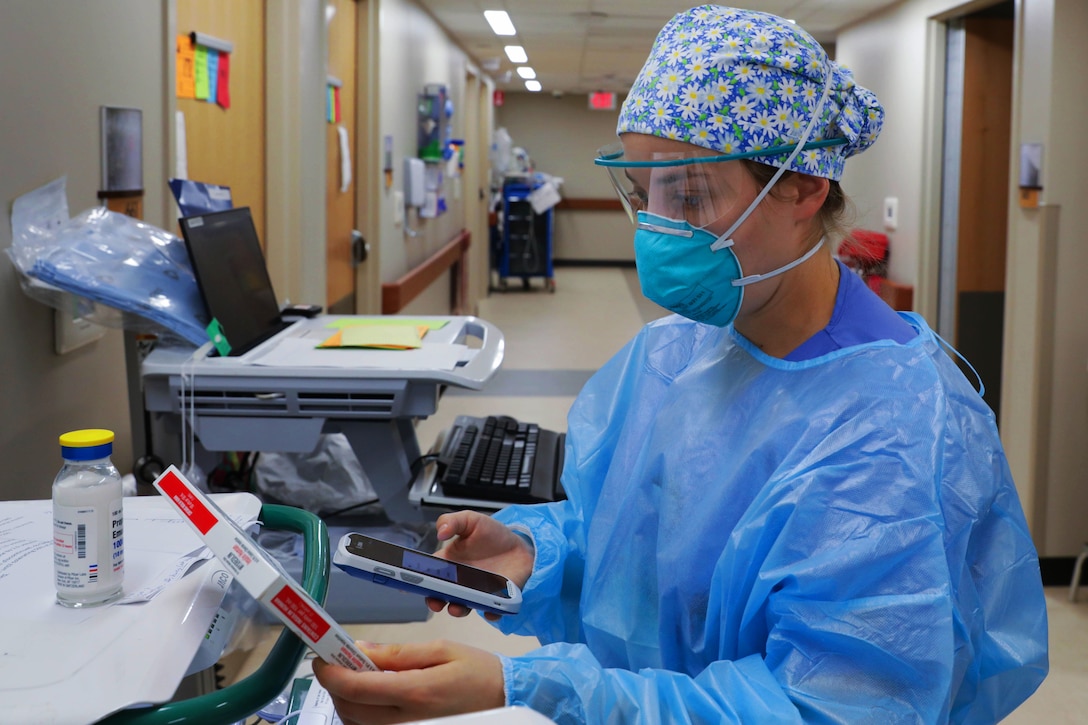 A sailor wearing a face mask, gloves and medical gown holds a patient's medication while checking it.