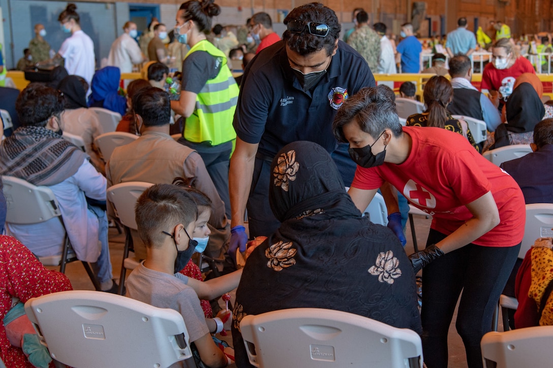 Volunteers pass out food and drinks to civilians sitting in a large room.