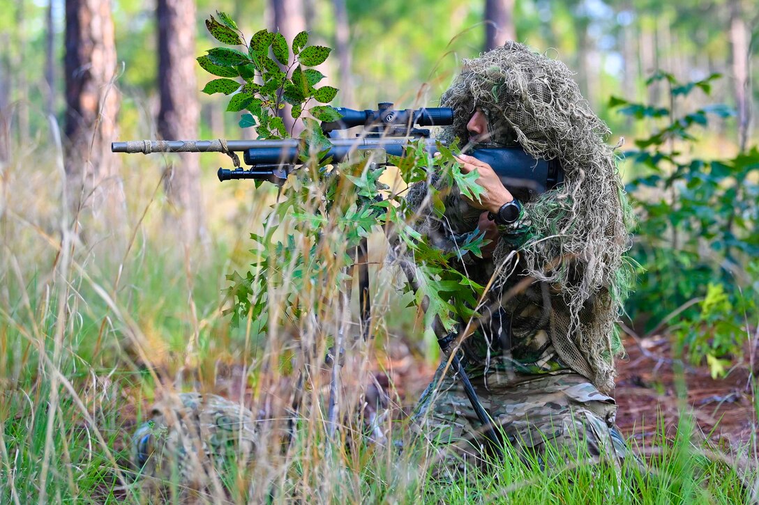 A person in camouflage looks through the scope of a weapon in a wooded area.
