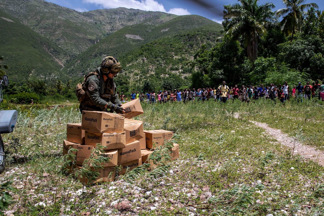 A sailor stands in a field with a stack of cardboard boxes as civilians walk toward him.