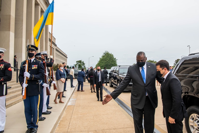 A man extends an arm out to show another man the way. Military honor guard members greet them.