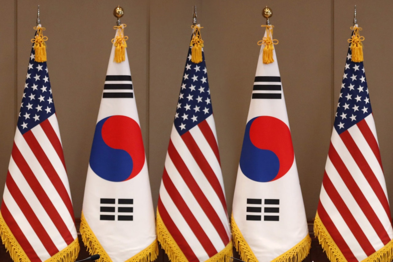 Three U.S. flags with two South Korean flags between them stand in a row against a wall.