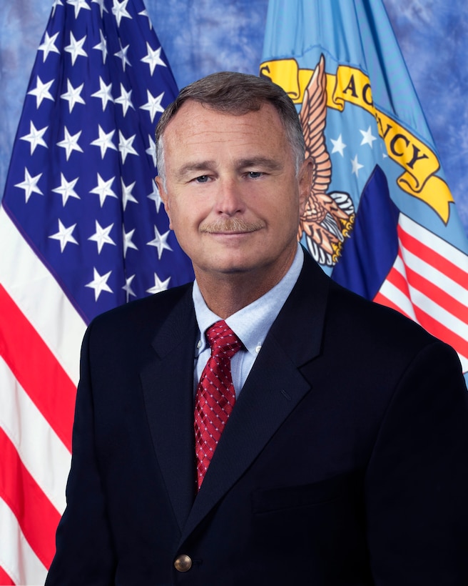 official photo man in black suit, red tie in front of the American and DLA flags.