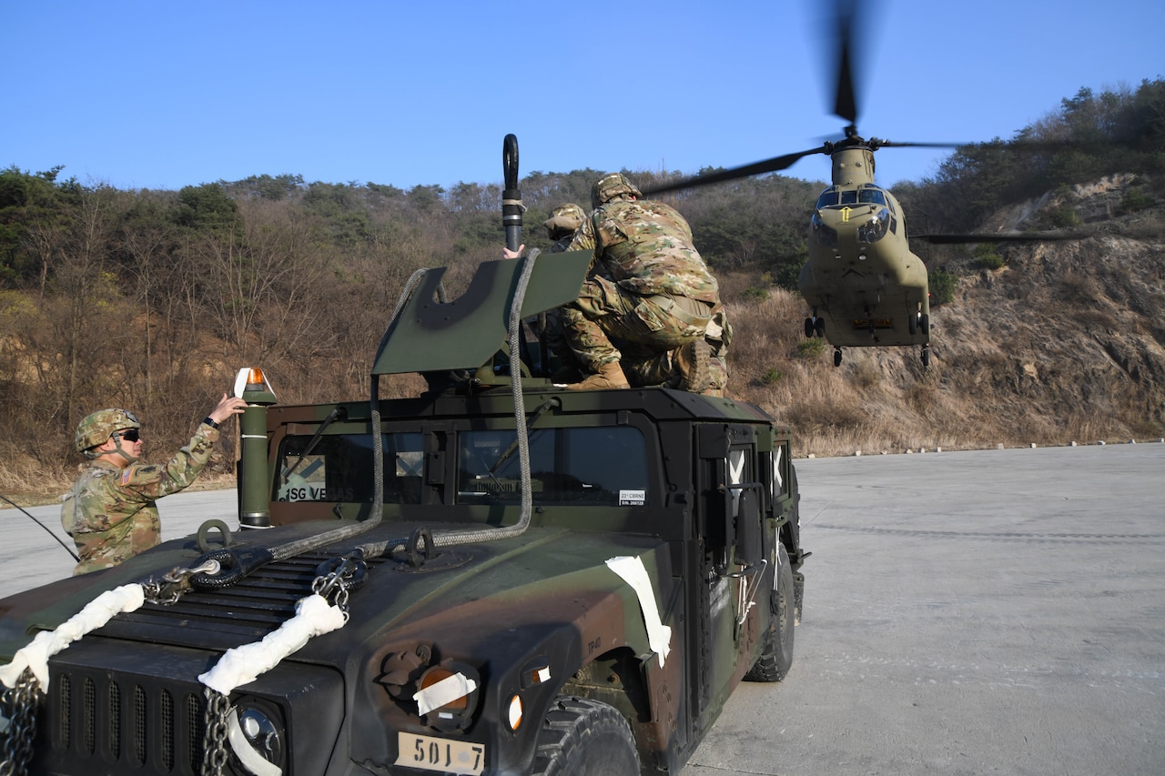 Some soldiers sit on or stand near a truck. A helicopter approaches.