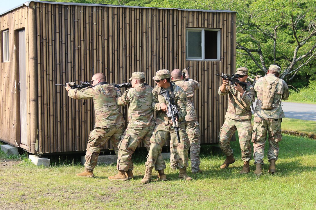 Soldiers with rifles stand outside a small, bamboo-covered building.