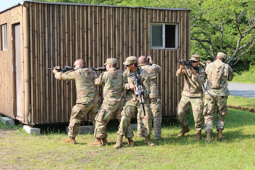 Soldiers with rifles stand outside a small, bamboo-covered building.