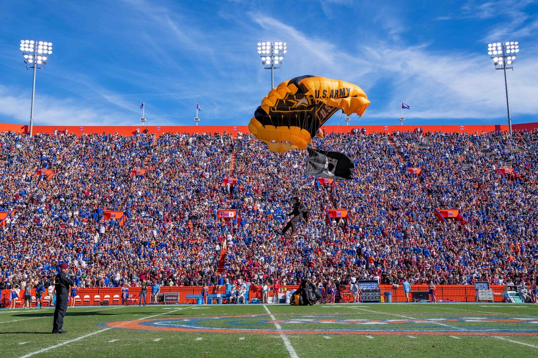 A soldier prepares to land at a stadium full of spectators, with a parachute and flag.