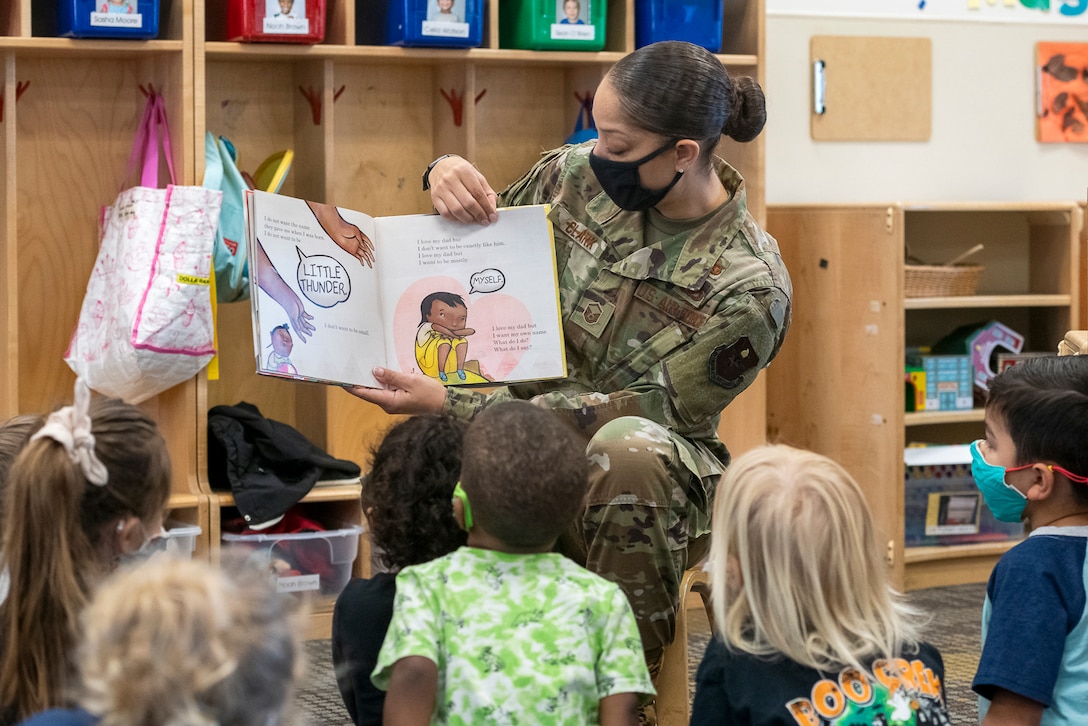 An airman in a chair holds up an illustrated book as kids sitting on the floor look at it.